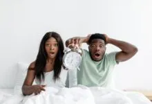 A surprised couple holding an alarm clock while sitting in bed, expressing shock.