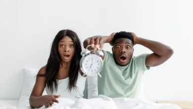 A surprised couple holding an alarm clock while sitting in bed, expressing shock.