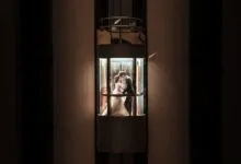 A newlywed couple shares a romantic kiss inside an illuminated glass elevator, embracing love and unity.