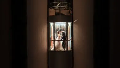 A newlywed couple shares a romantic kiss inside an illuminated glass elevator, embracing love and unity.