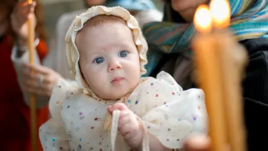 A baby in a bonnet during a baptism ceremony, surrounded by candles and family.