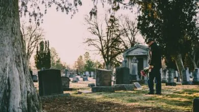 A person stands in a peaceful cemetery holding flowers in remembrance.