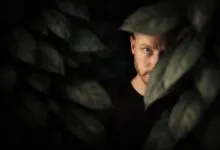 A man with one eye visible peers through thick leaves, creating a mysterious and intriguing effect.