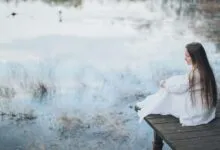 Side view of peaceful young female wearing elegant dress sitting on wooden boardwalk near lake