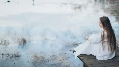 Side view of peaceful young female wearing elegant dress sitting on wooden boardwalk near lake