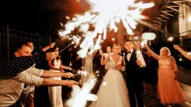 Happy young bride and groom standing on street near unrecognizable guests with burning sparklers during wedding party in evening