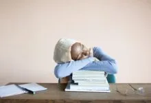 Sleeping African American female student in headscarf sitting at table with opened notebook and lying on stack of textbooks