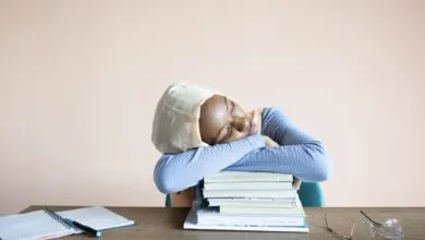 Sleeping African American female student in headscarf sitting at table with opened notebook and lying on stack of textbooks