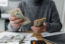 Close-up of person counting cash with a calculator and paperwork on a desk.