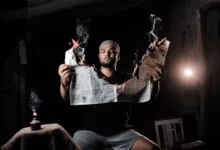 A man holds a burning newspaper, creating a dramatic and conceptual visual in a dimly lit room.