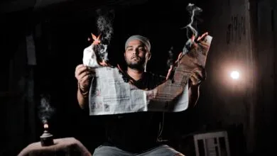 A man holds a burning newspaper, creating a dramatic and conceptual visual in a dimly lit room.