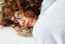 Close-up of a woman with curly hair and glasses resting her head on US dollar bills, symbolizing dreams of wealth.