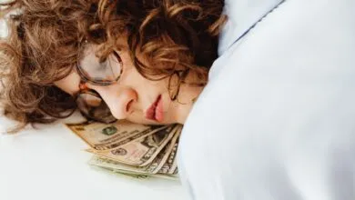 Close-up of a woman with curly hair and glasses resting her head on US dollar bills, symbolizing dreams of wealth.