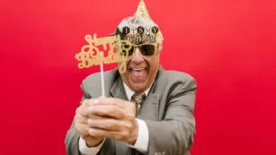 Elderly man in a suit celebrating his birthday with festive props and a red backdrop.