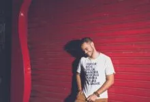 Casually dressed young man posing playfully against a vibrant red steel shutter with text t-shirt design.