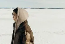 Жінка в утепленій куртці з капюшоном поверх худі стоїть на засніженому відкритому березі